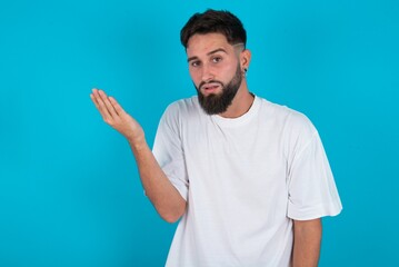 Fototapeta premium Studio shot of frustrated bearded caucasian man wearing white T-shirt over blue background gesturing with raised palm, frowning, being displeased and confused with dumb question.