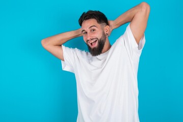 Fototapeta na wymiar bearded caucasian man wearing white T-shirt over blue background relaxing and stretching, arms and hands behind head and neck smiling happy