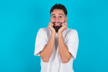Portrait of bearded caucasian man wearing white T-shirt over blue background being overwhelmed, expressing excitement and happiness with closed eyes and hands near face.
