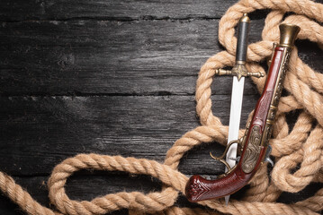 Pirate concept background. Musket gun, dagger and mooring rope on the black wooden table flat lay...