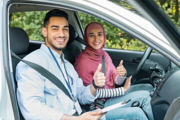 Smiling millennial arab female and male teacher in hijab showing thumbs up in car, approving driving lesson