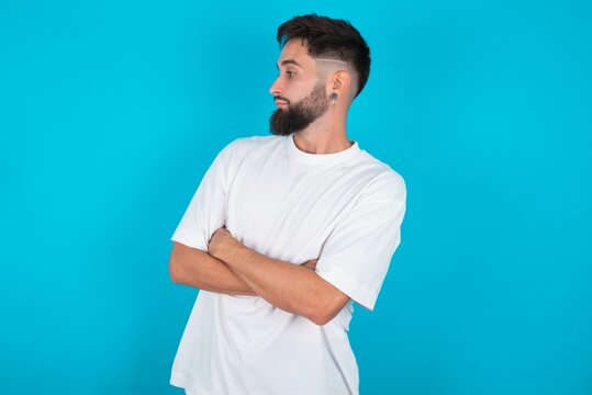 Image of upset bearded caucasian man wearing white T-shirt over blue background with arms crossed. Looking with disappointed expression aside after listening to bad news.