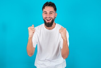 Fototapeta premium Portrait of bearded caucasian man wearing white T-shirt over blue background looks with excitement at camera, keeps hands raised over head, notices something unexpected reacts on sudden news.