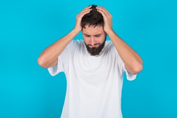 Fototapeta na wymiar bearded caucasian man wearing white T-shirt over blue background holding head with hands, suffering from severe headache, pressing fingers to temples