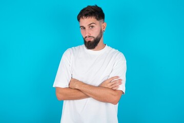 bearded caucasian man wearing white T-shirt over blue background frowning his face in displeasure,...