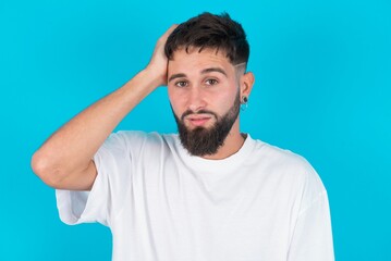 Obraz premium Portrait of confused bearded caucasian man wearing white T-shirt over blue background holding hand on hair and frowning, panicking, losing memory. Worried and anxious can not remember anything.