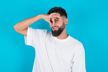 Displeased bearded caucasian man wearing white T-shirt over blue background plugs nose as smells something stink and unpleasant, feels aversion, hates disgusting scent.