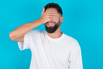 Happy bearded caucasian man wearing white T-shirt over blue background closing eyes with hand going to see surprise prepared by friend standing and smiling in anticipation for something wonderful.