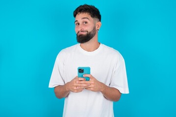 Happy bearded caucasian man wearing white T-shirt over blue background listening to music with earphones using mobile phone.