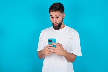 bearded caucasian man wearing white T-shirt over blue background looks with bugged eyes, holds modern smart phone, receives unexpected message from friend, reads reminder.