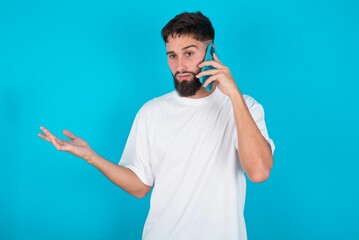 bearded caucasian man wearing white T-shirt over blue wall talking on the phone stressed with hand on face, shocked with shame and surprise face, angry and frustrated. Fear and upset for mistake.