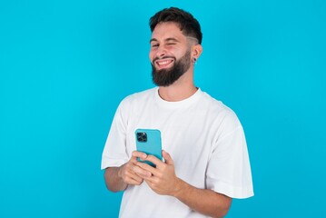 Pleased bearded caucasian man wearing white T-shirt over blue background using self phone and looking and winking at the camera. Flirt and coquettish concept.