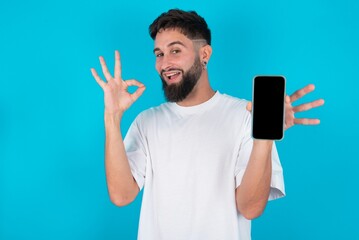 Excited bearded caucasian man wearing white T-shirt over blue background showing smartphone blank screen, blinking eye and doing ok sign with hand.  Advertisement concept.