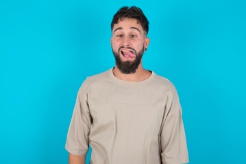 Funny bearded caucasian man wearing casual T-shirt over blue background makes grimace and crosses eyes plays fool has fun alone sticks out tongue.