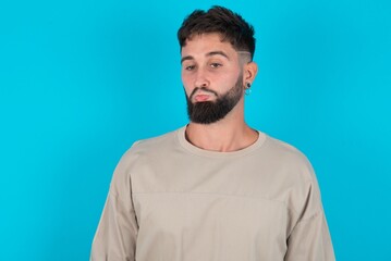 Dissatisfied bearded caucasian man wearing casual T-shirt over blue background purses lips and has unhappy expression looks away stands offended. Depressed frustrated model.