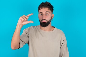 bearded caucasian man wearing casual T-shirt over blue background purses lip and gestures with hand, shows something very little.