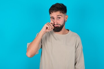 Disappointed dejected bearded caucasian man wearing casual T-shirt over blue background wipes tears stands stressed with gloomy expression. Negative emotion
