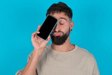 Adorable bearded caucasian man wearing casual T-shirt over blue background holding modern device covering eye with lips pouted