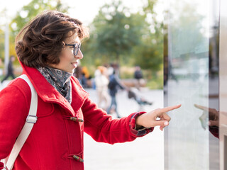Curly woman in red coat is scrolling screen on outdoor electronic navigation device. Modern electronic panels on streets for e-commerce or touristic maps.