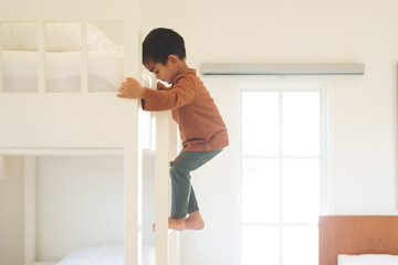 Fototapeta na wymiar Asian toddler boy is trying to climb up the ladder of bunk bed in the bedroom. Concept of childhood, kid development and physical activity.
