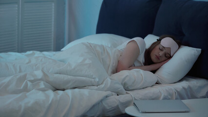 Woman in mask sleeping on bed near laptop on bedside table.