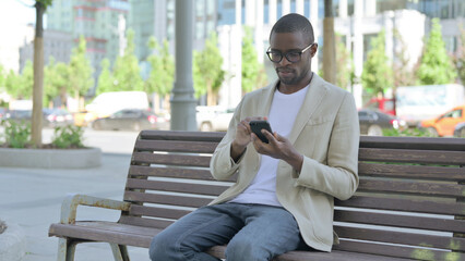 Fototapeta premium African Man Browsing Internet on Smartphone while Sitting Outdoor on Bench