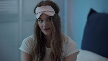 Blonde woman in sleep mask looking at camera in bedroom in evening.