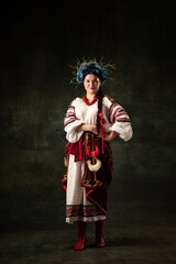 Adorable woman wearing national folk Ukrainian attire posing isolated over dark vintage background. Fashion, beauty, cultural heritage, eras comparison