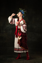 Enjoy. Creative portrait of beautiful Ukrainian woman wearing traditional folk costume tasting varenik isolated over dark background.