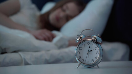 Alarm clock on bedside table near blurred woman sleeping on bed.