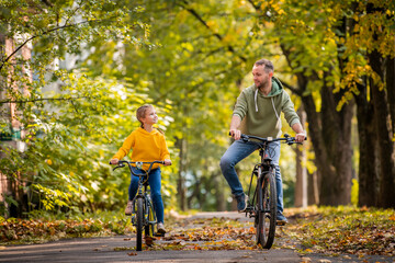 Happy father and daughter ride bicycles in autumn park on sunny day.