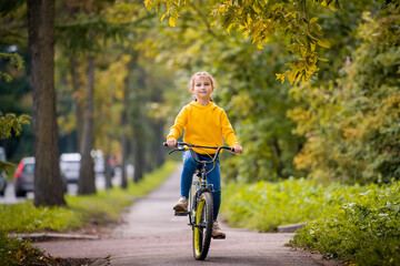 smiling girl of ten in a yellow sweater rides bicycle on autumn street under the branches of a red...