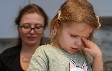 Sleepy, moody little girl stands in kitchen and rubs eyes while her mom is on phone on social networks. Mother plays on phone but does not play with her daughter