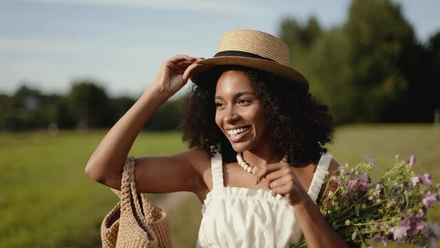 4k Portrait of attractive african-american woman in hat and white clothes with bag of flowers walking in the summer field. Countryside and farmland style.