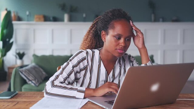 Young attentive African American woman looking at laptop screen touching face and reading article or studying at online university dressed in business casual style sits at table in own house