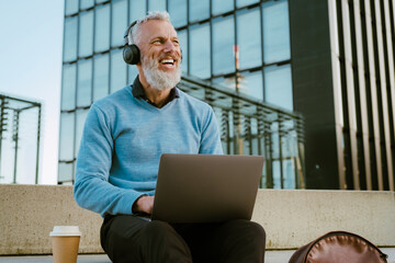 Mature grey man listening music while working with laptop at street
