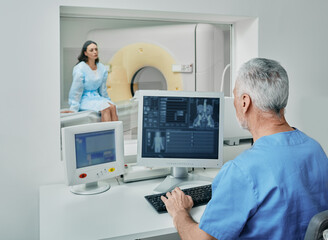 Radiologist before CT scan of female patient's body using CT scanner from control room behind...