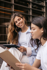 Two cute young caucasian classmates spend leisure time together outdoors. Blonde and brunette wear...