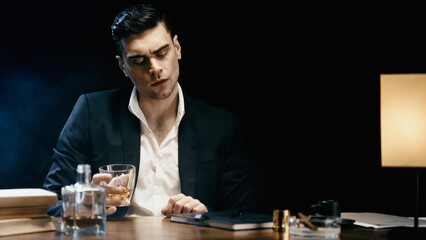 Businessman holding glass of whiskey near books and lamp on black.