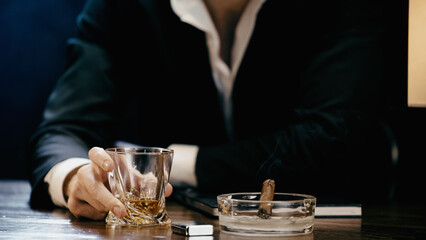 Cropped view of businessman holding glass of whiskey near cigar and lighter isolated on black.