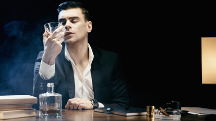 Businessman holding glass of whiskey near books and cigar in ashtray on black with smoke.