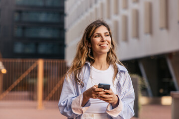Fototapeta premium Beautiful young caucasian girl uses smartphone spending leisure time on street. Blonde smiles looking away, wears casual clothes. City life concept