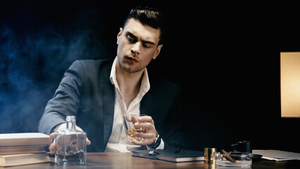 Businessman holding glass of whiskey near books and cigar on black background with smoke.