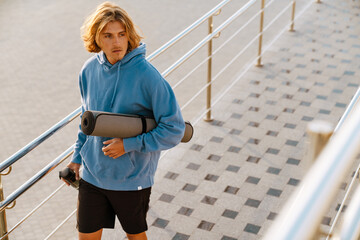 Young blonde man walking with mat and water bottle after workout