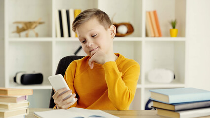 pensive schoolboy using smartphone near notebook and books on desk.