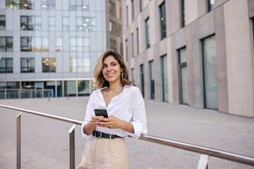 Stylish young caucasian girl works with mobile phone on street with office buildings. Blonde looks to side, wears classic style of clothing. Management concept