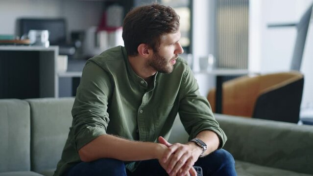 nervous man is waiting mobile call or message, looking at smartphone in hands, sitting alone in living room