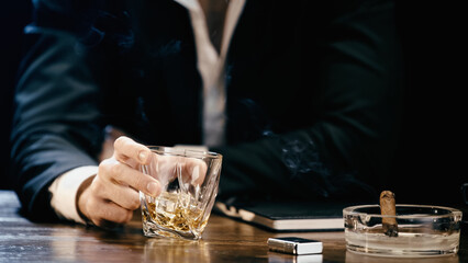 Cropped view of blurred businessman holding glass of whiskey near cigar in ashtray isolated on black. - Powered by Adobe