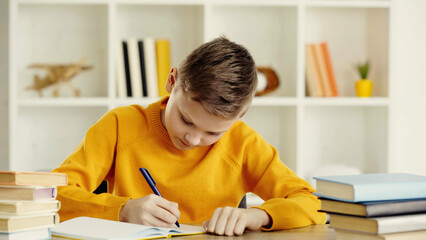preteen boy writing in notebook while doing homework near books on wooden table at home.