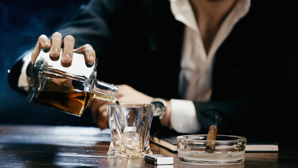 Cropped view of blurred businessman pouring whiskey near cigar in ashtray on black.